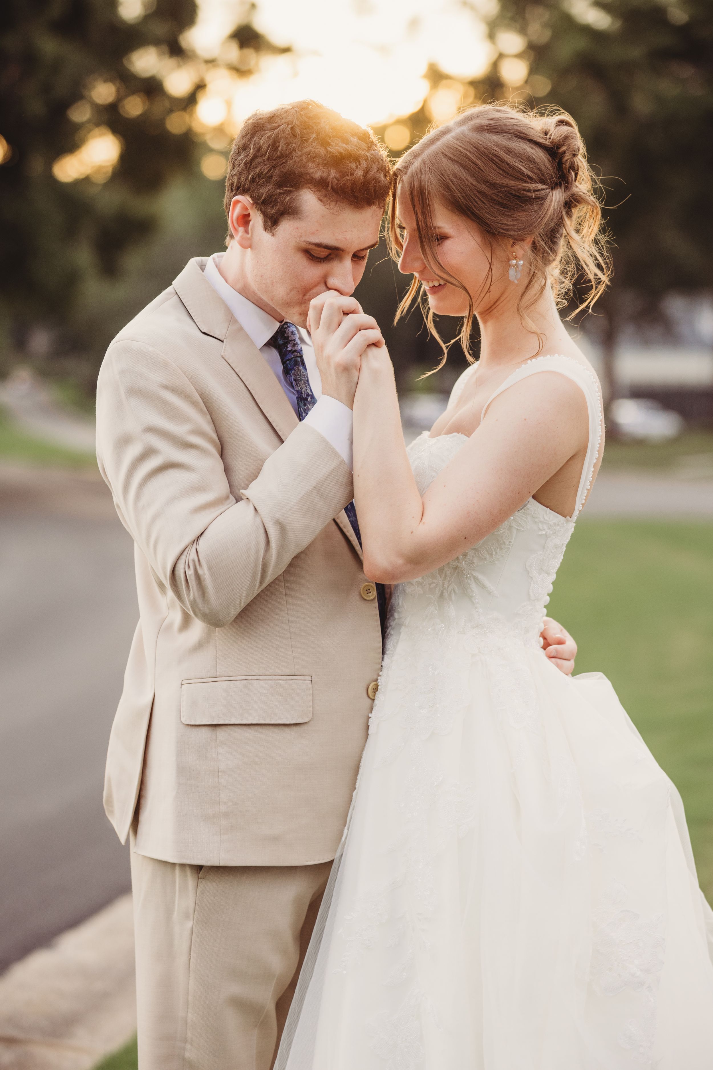 Groom in beige suit kissing bride's hand at golden hour outdoor wedding ceremony.