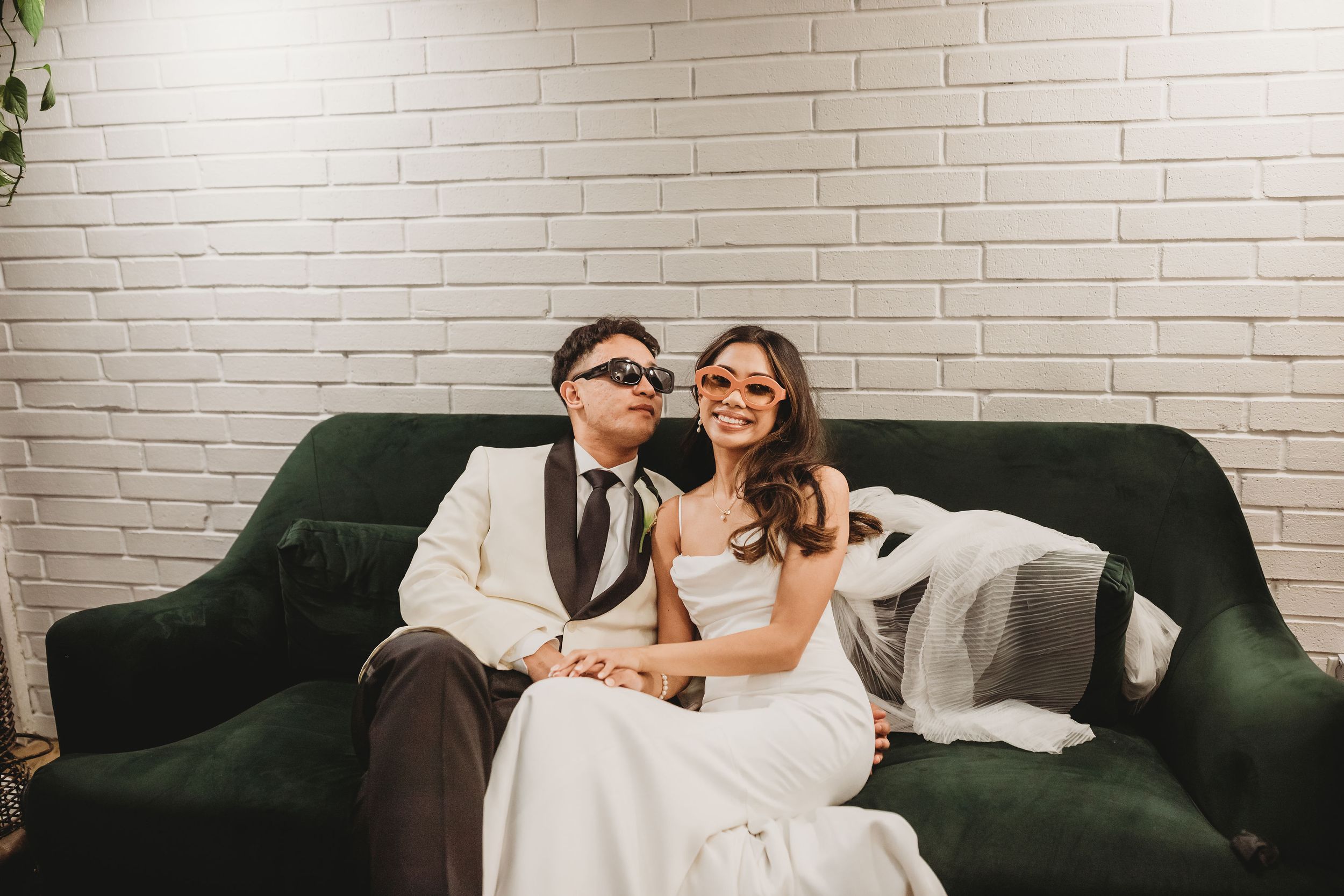 Bride and groom sitting on green velvet sofa wearing fun oversized sunglasses, smiling together against white brick wall.