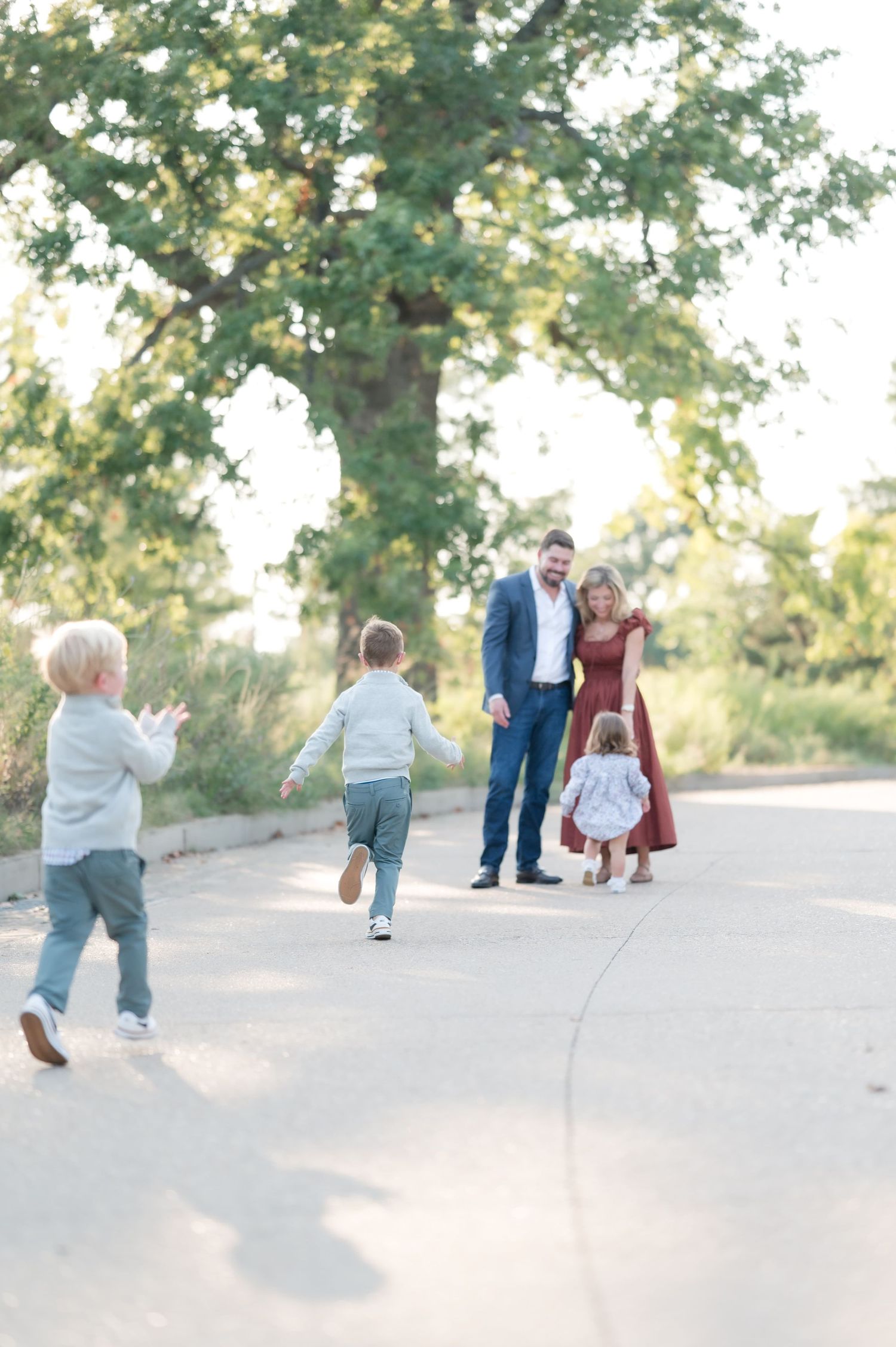 A family of three poses outdoors, parents in blue and rust dress admiring their toddler girl on a tree-lined path.