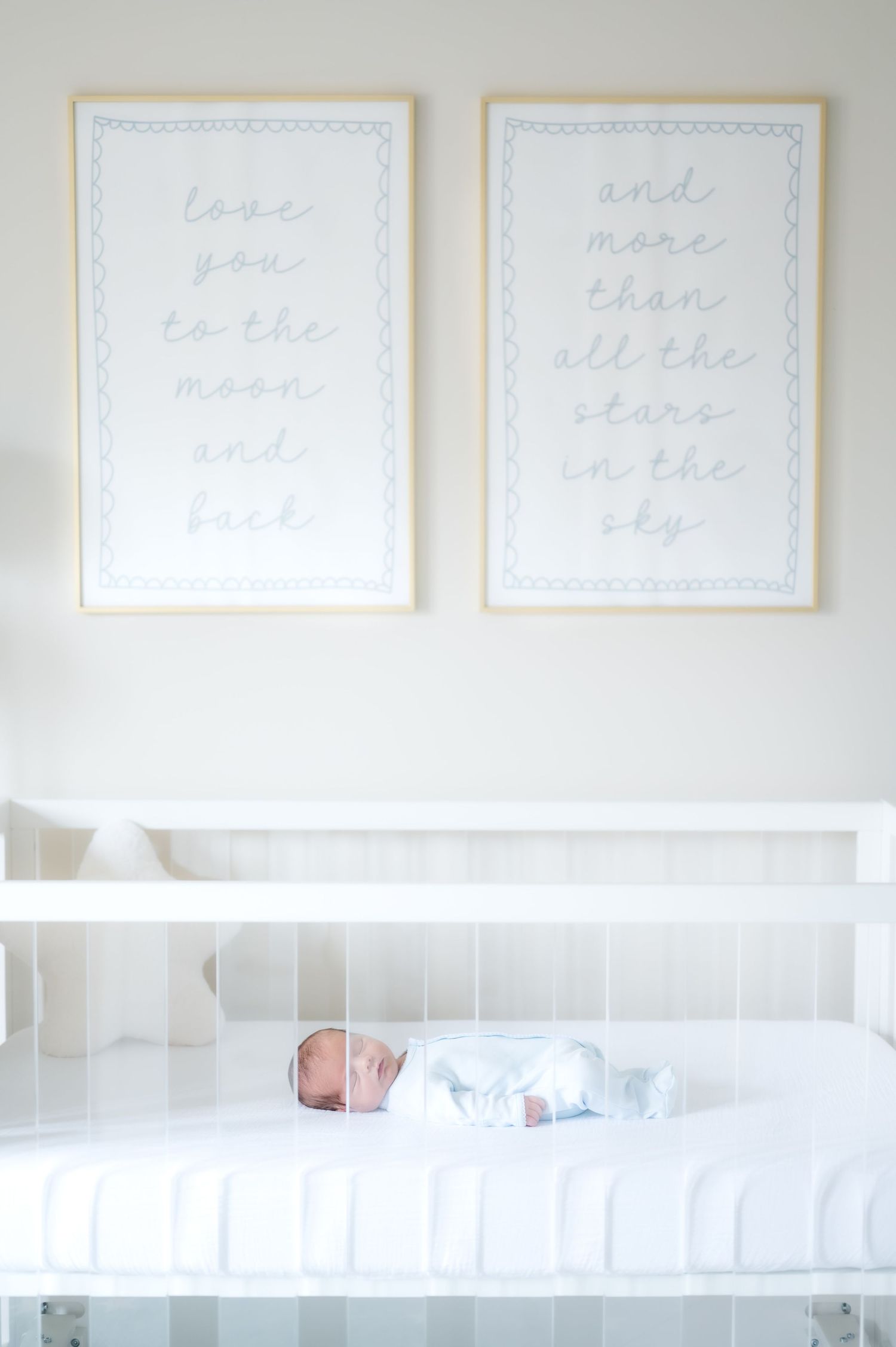 Newborn baby sleeping peacefully in a white crib beneath two framed nursery prints with sweet love quotes.