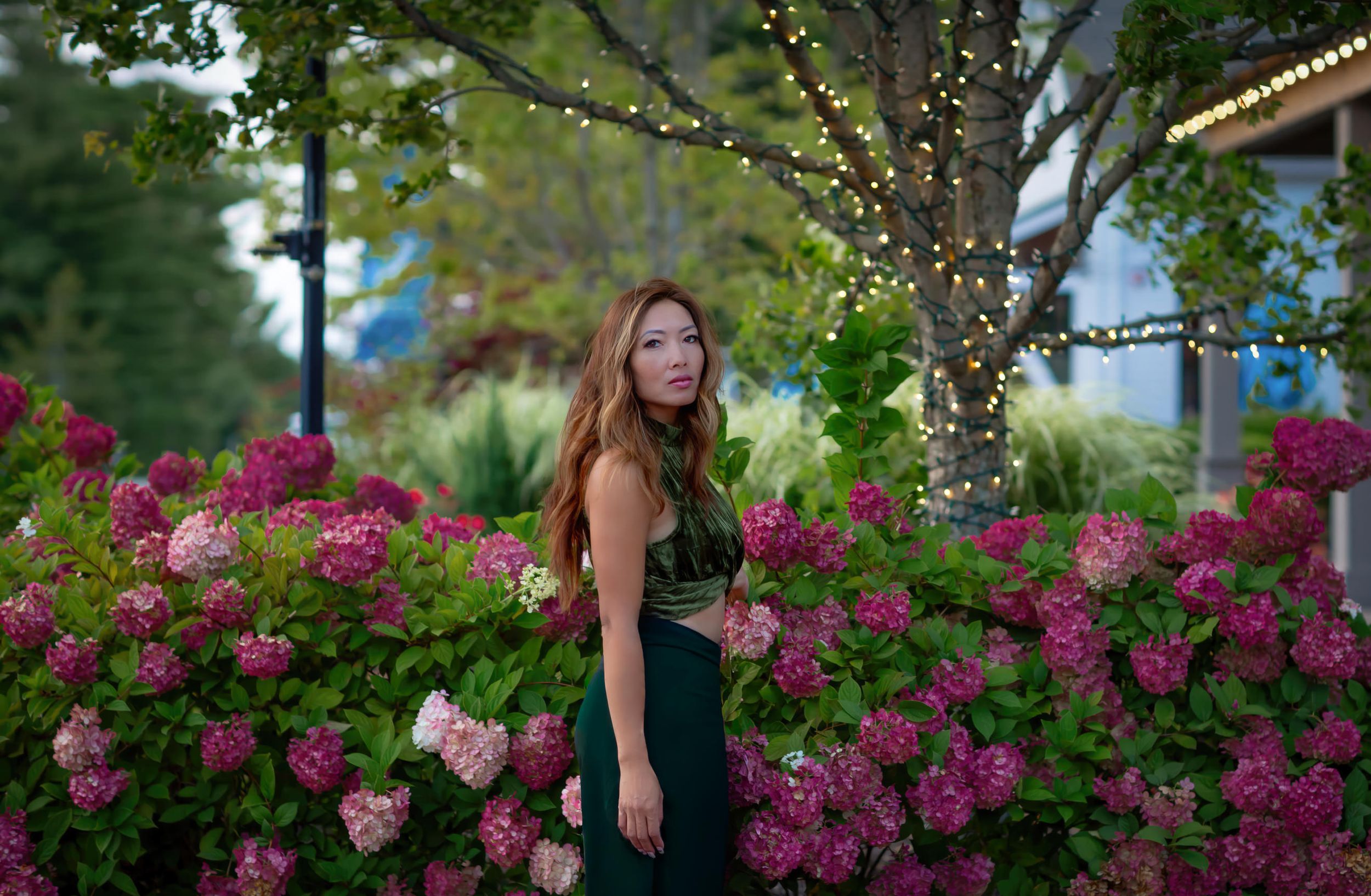 Woman in green dress poses among pink hydrangeas with string lights and trees at dusk.
