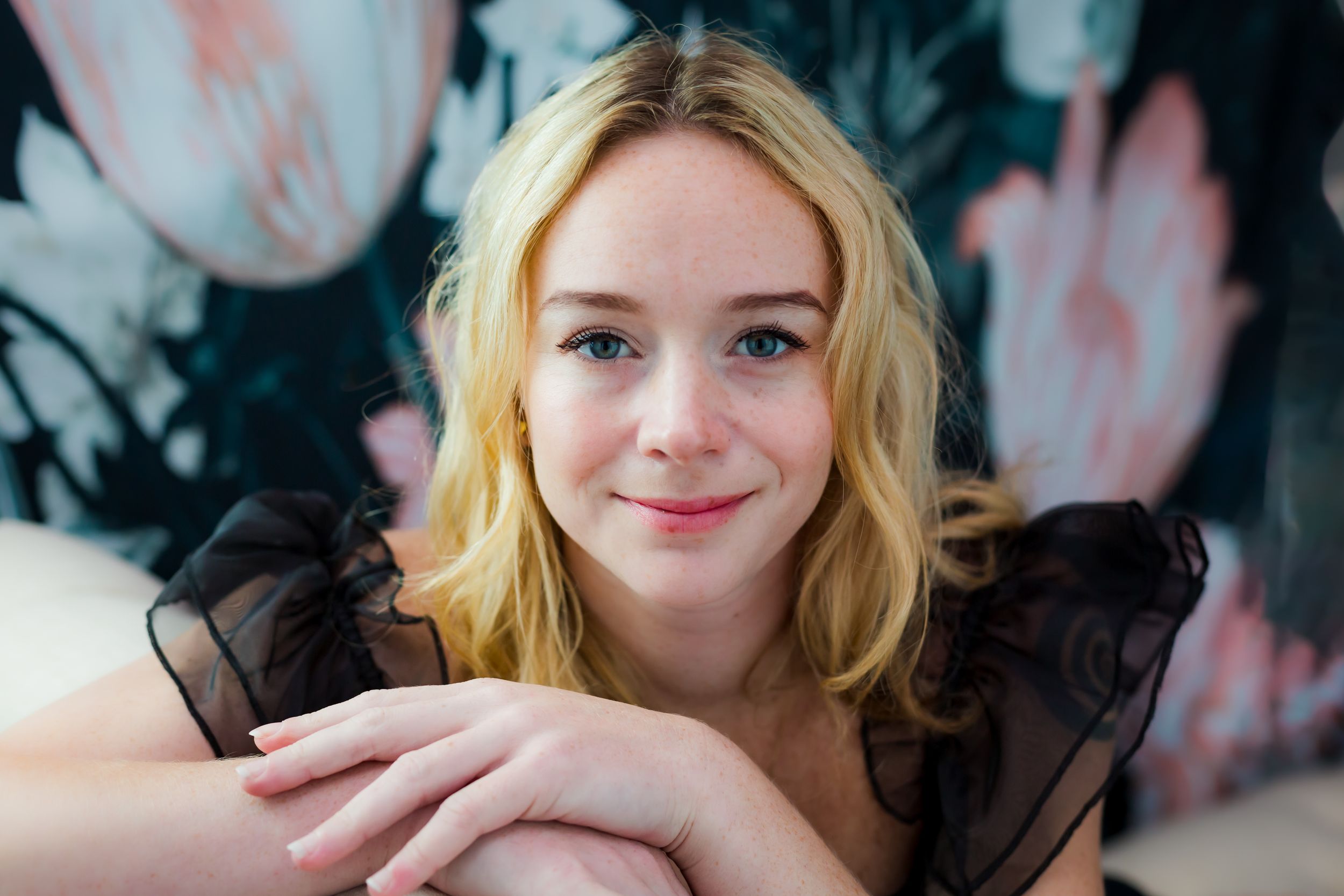 Young blonde woman with a warm smile resting her chin on clasped hands against a floral background.