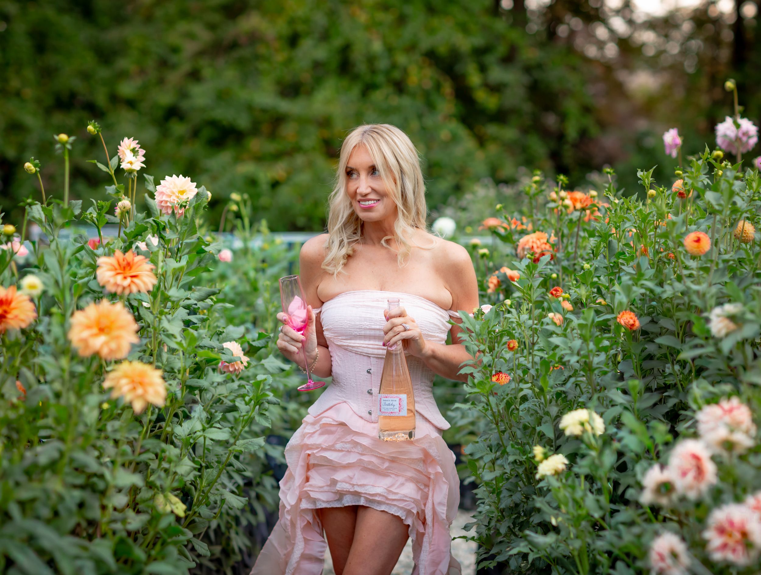 Blonde woman in pink off-shoulder dress holding perfume bottle surrounded by colorful dahlia flowers in a lush garden.