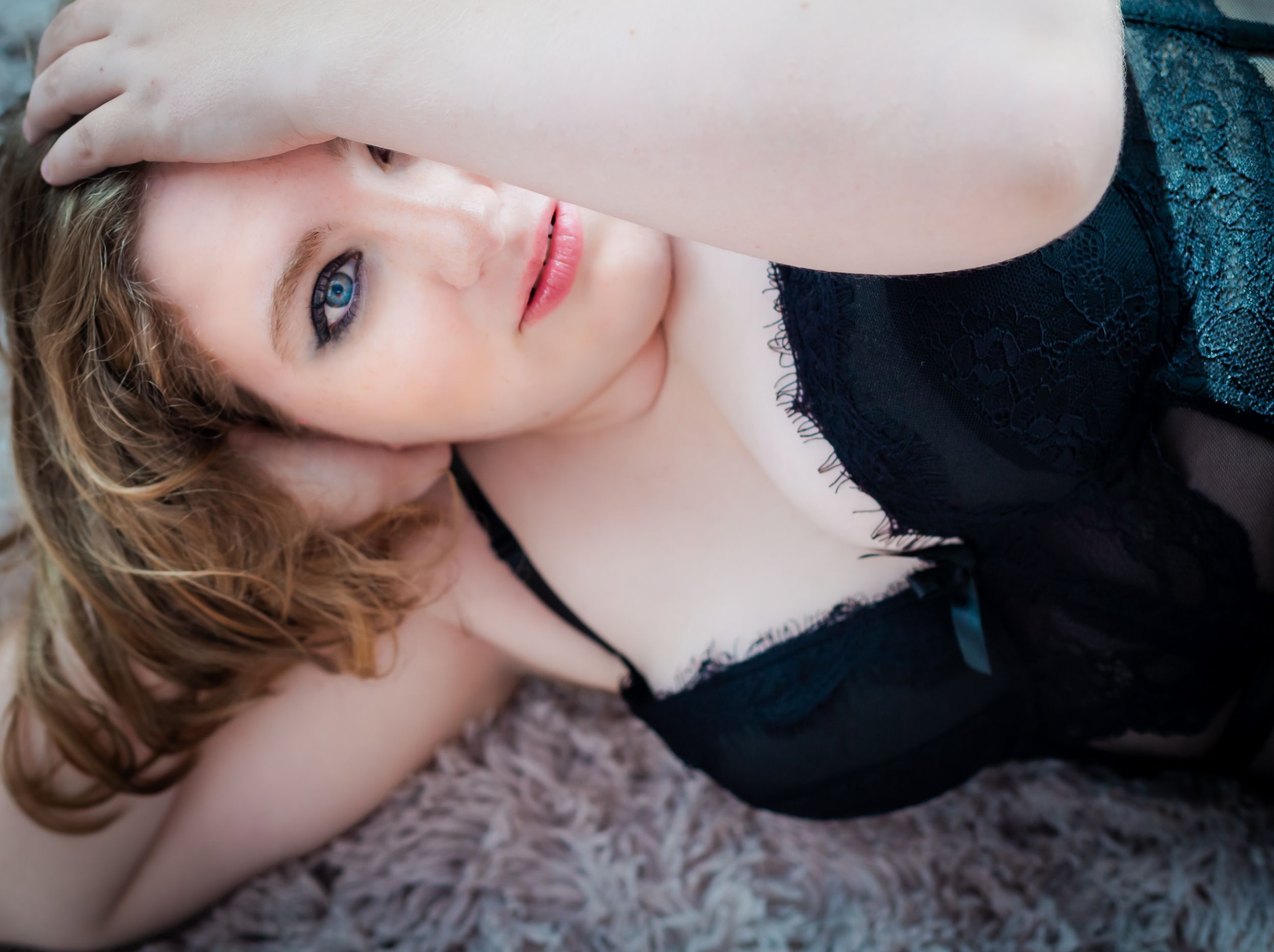 Young woman in black lace lingerie posing thoughtfully on a soft rug, looking up with blue eyes.