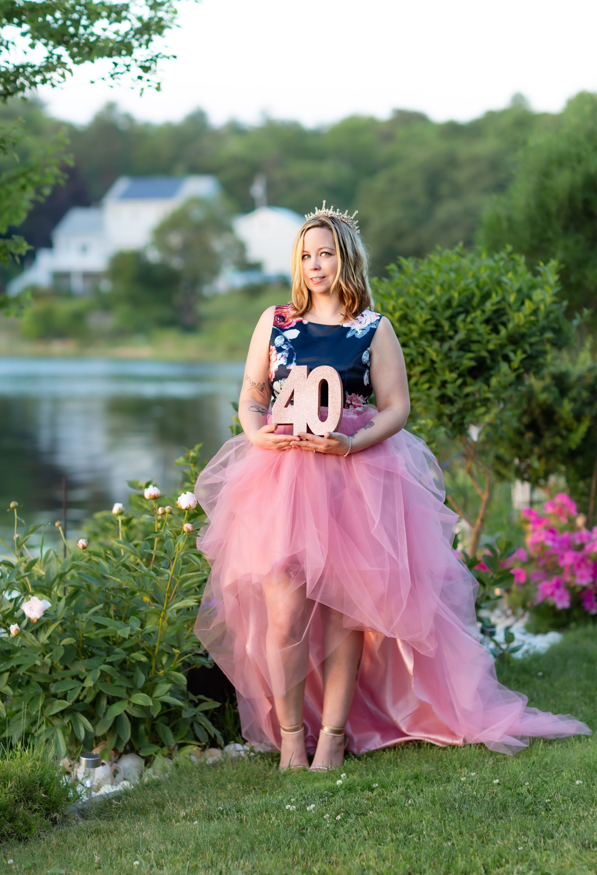 Woman in pink tulle skirt and floral top holds a 40 sign, wearing a tiara, posing in a garden near a lake.