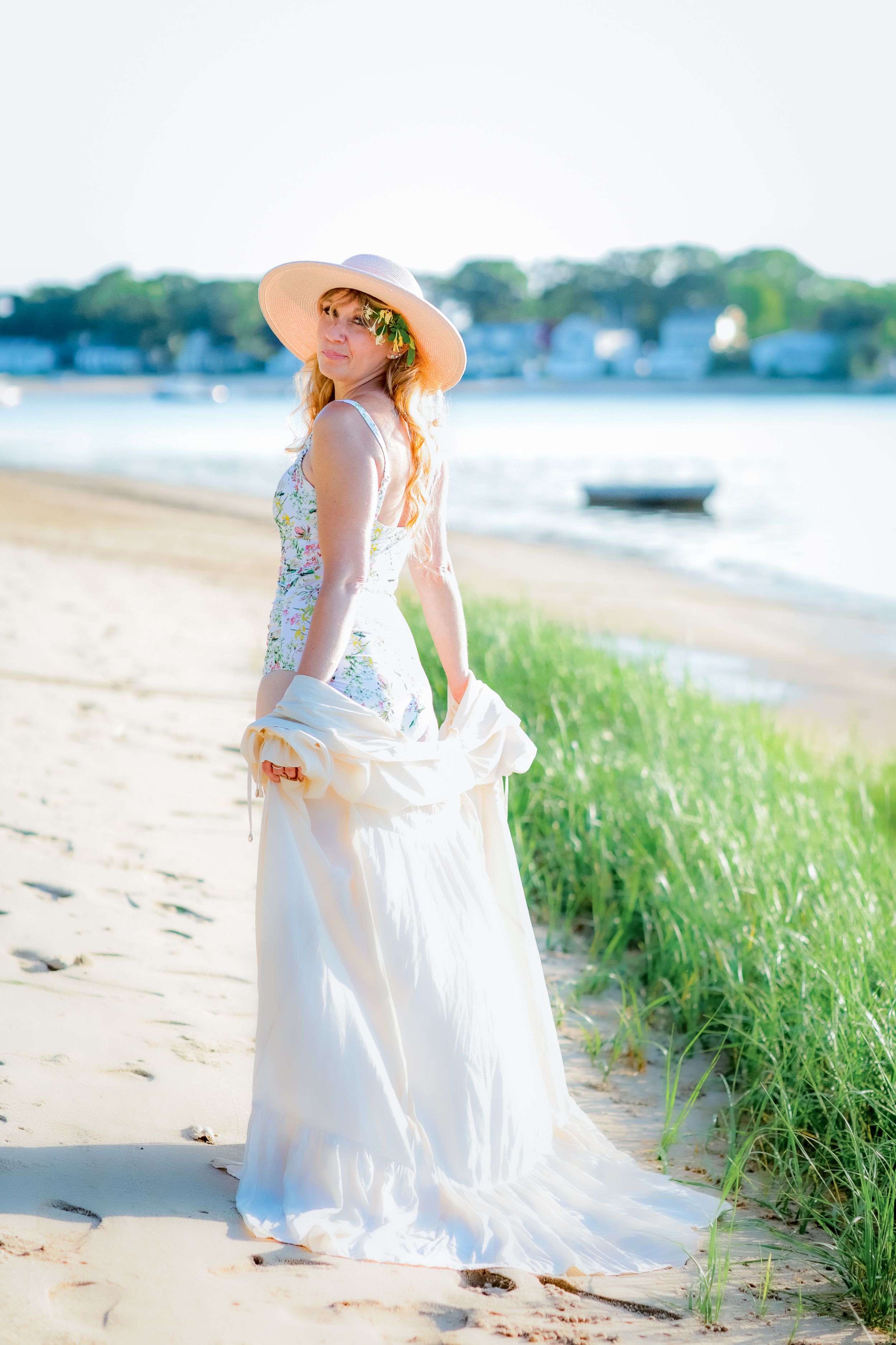 Woman in white flowy dress and sun hat standing on sandy beach near ocean grasses in golden hour light.
