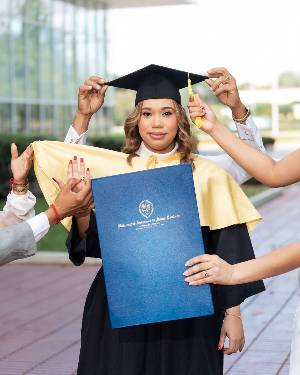 A smiling graduate in black cap and gown with gold hood holds a blue diploma cover while others adjust her mortarboard.