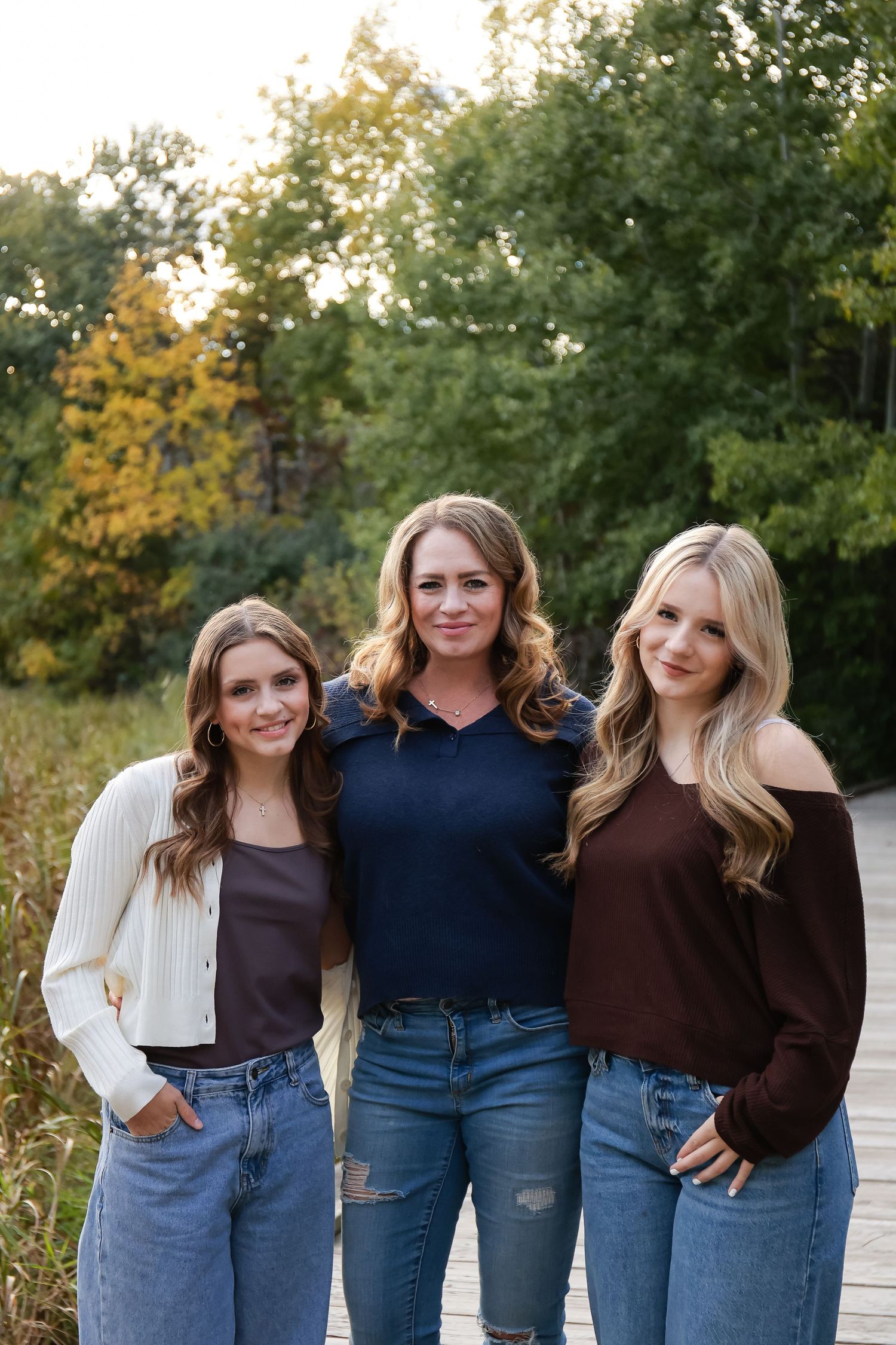 Three smiling women pose together on a wooden boardwalk surrounded by fall foliage.