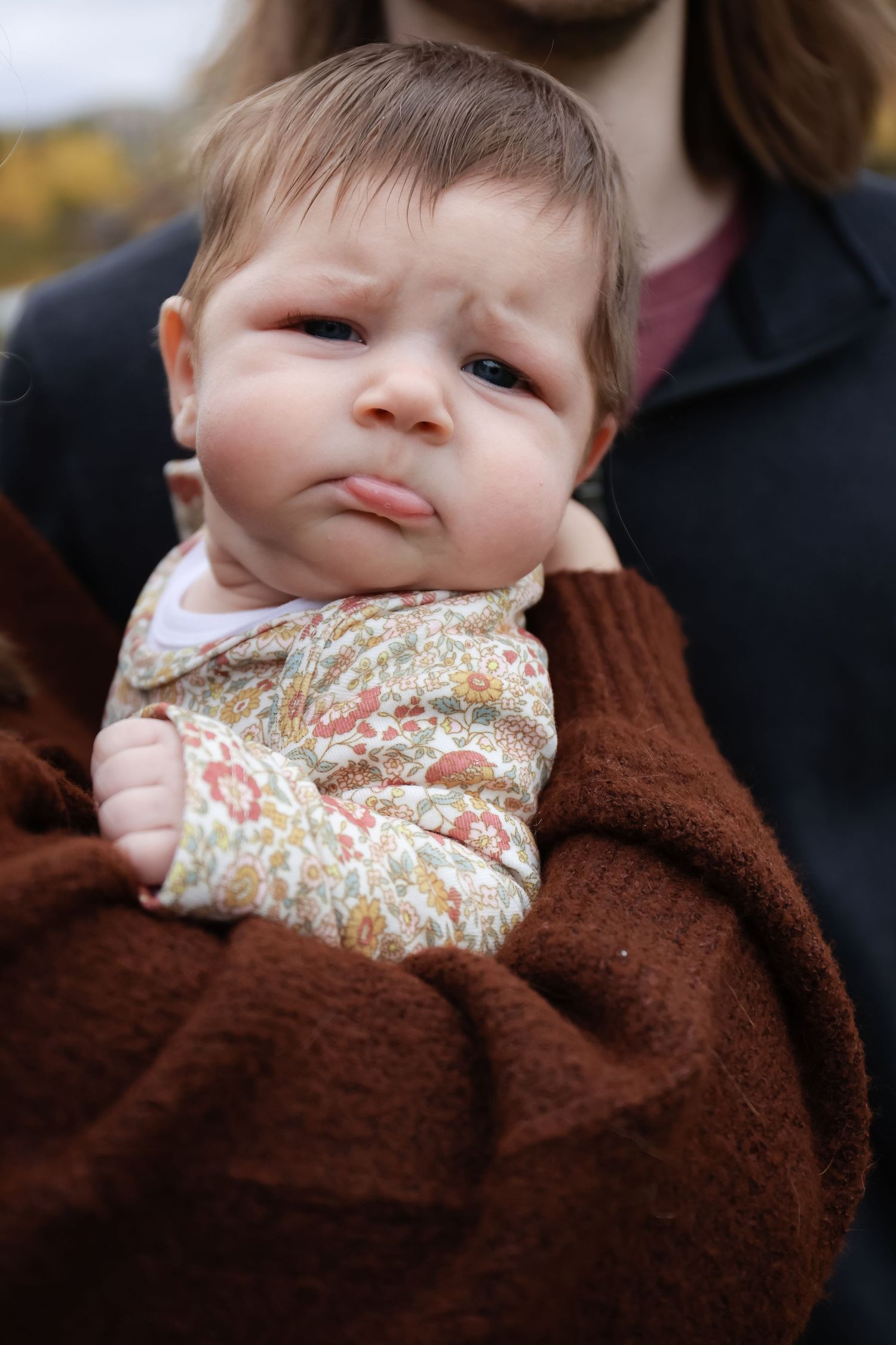 Close-up of newborn baby with pouty expression wrapped in brown blanket being held by parent outdoors.