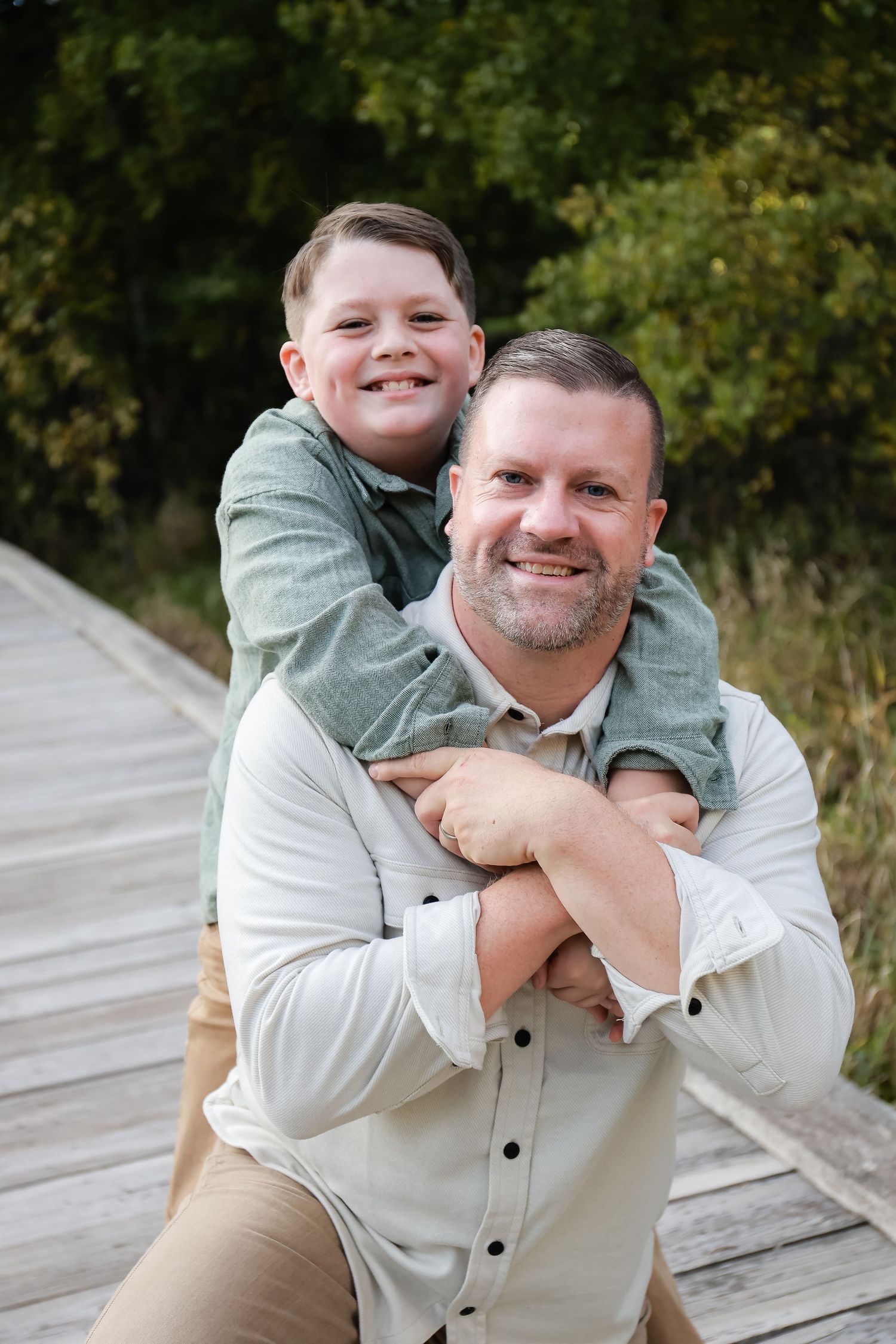 A smiling father gives his young son a piggyback ride on a wooden boardwalk.