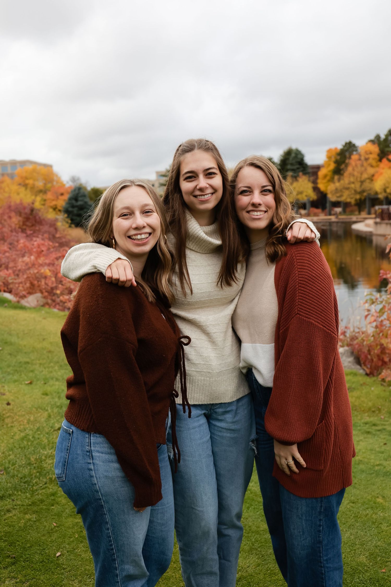 Three smiling young women in fall sweaters posing together near colorful autumn foliage by a pond.