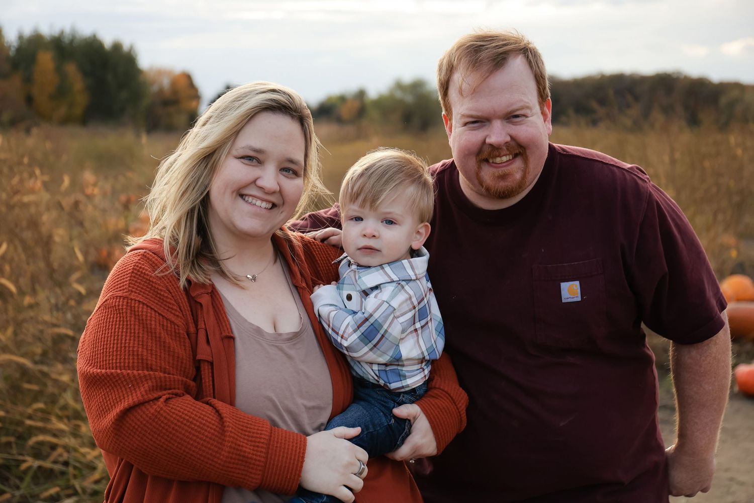 A happy young family with a baby poses at a pumpkin patch during autumn.