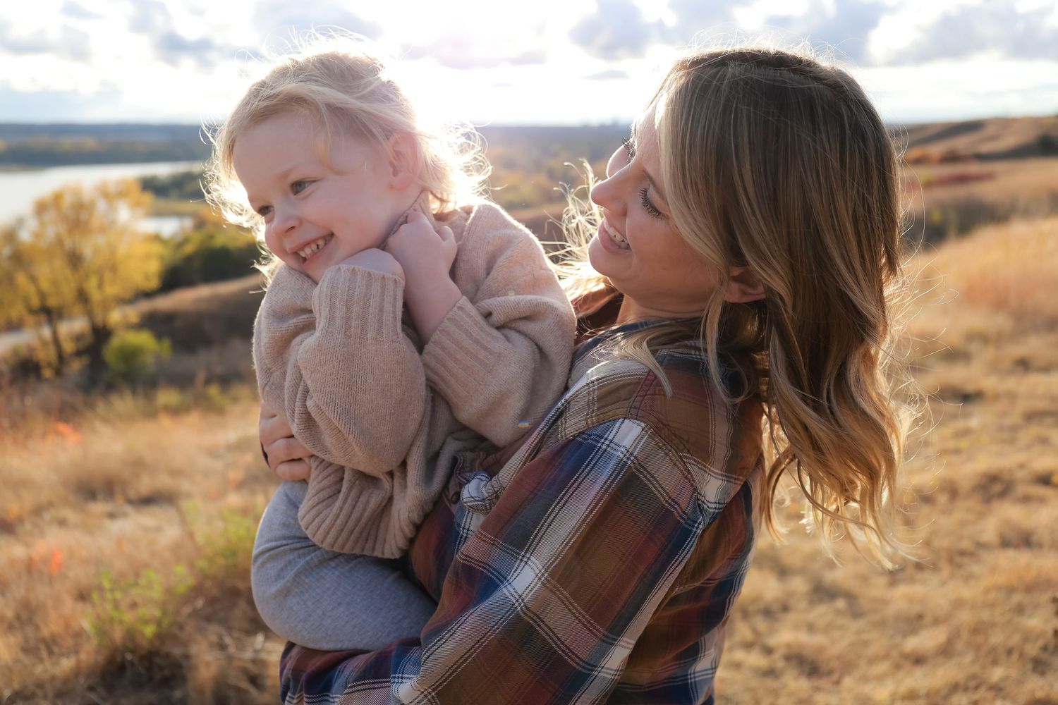 Mother in plaid shirt holding laughing blonde toddler in beige sweater in golden autumn field.