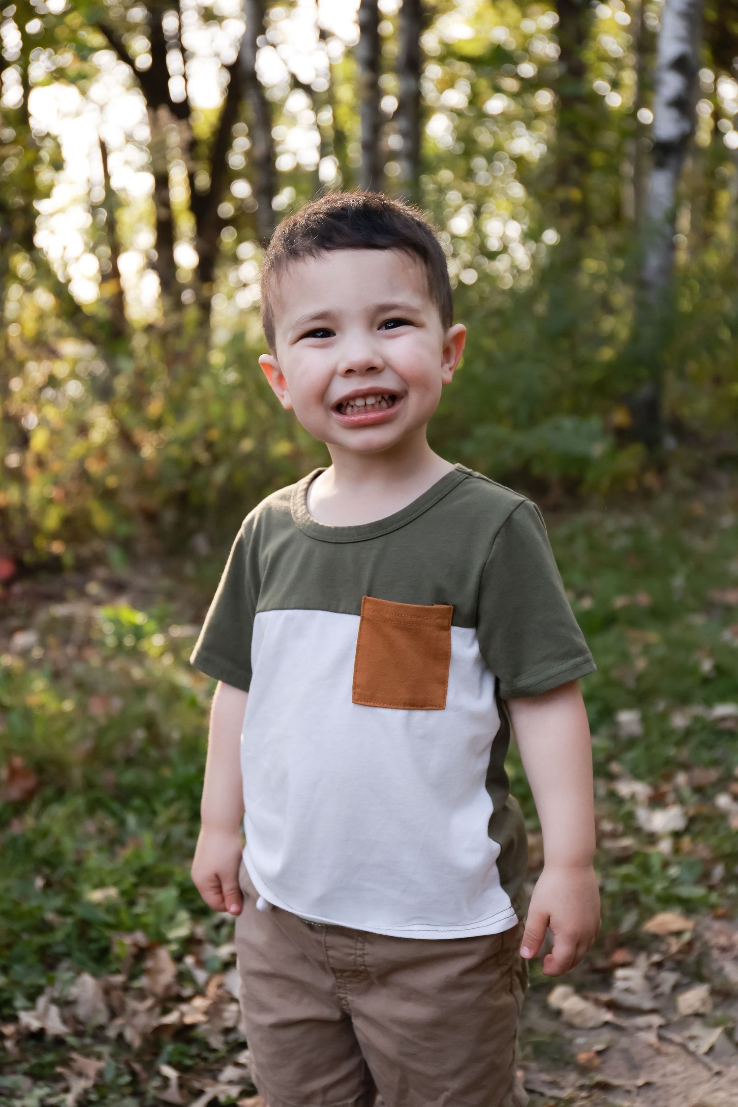 A cheerful young boy laughs while standing among autumn leaves in a forest.