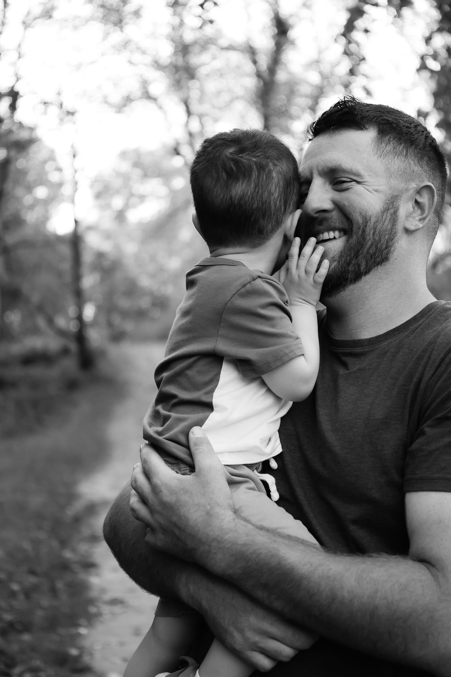 A bearded father laughs joyfully while holding his young child in a forest.