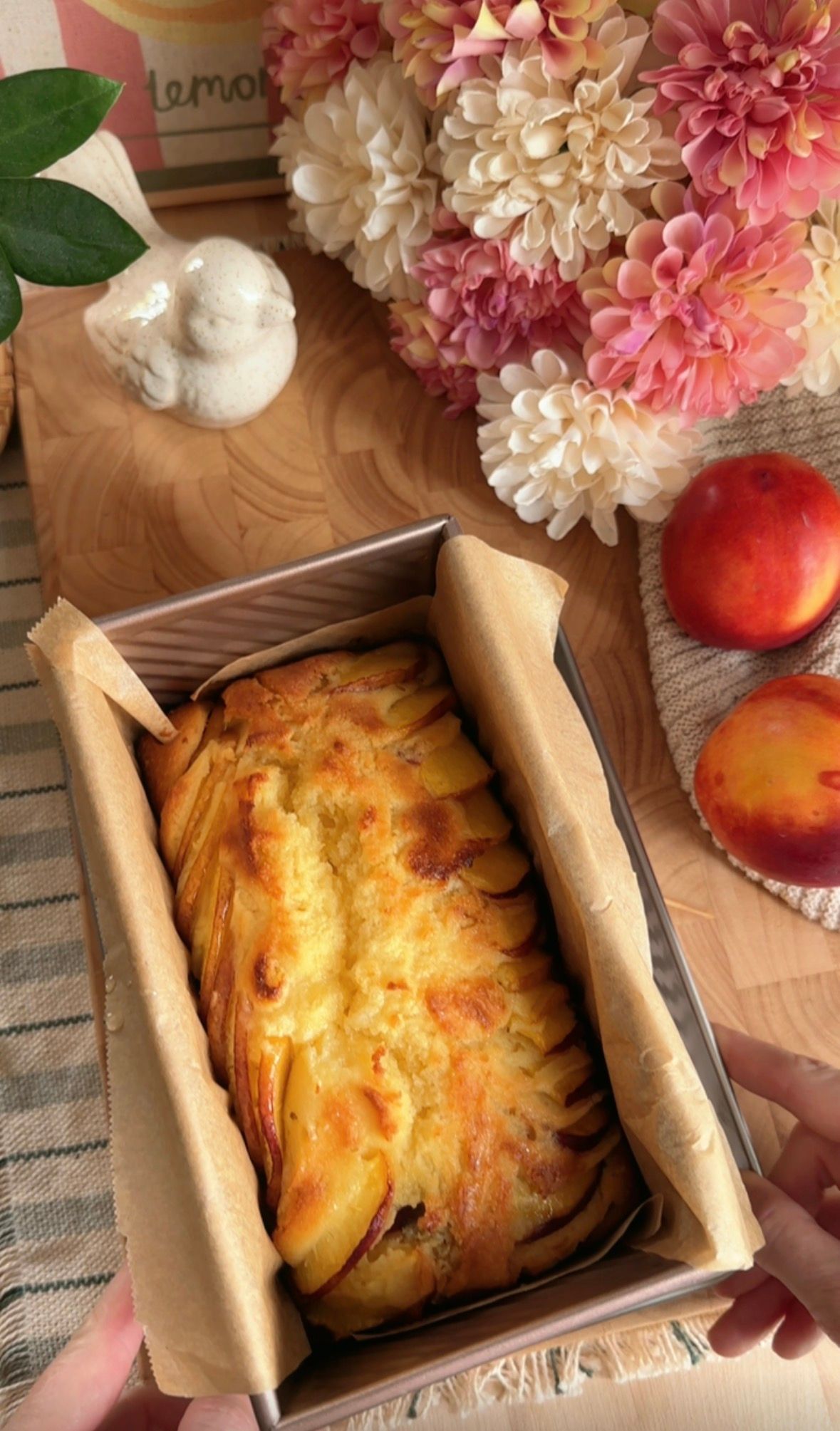Hands slicing a golden peach pull-apart bread loaf on a wooden board with fresh peaches and pink flowers nearby.