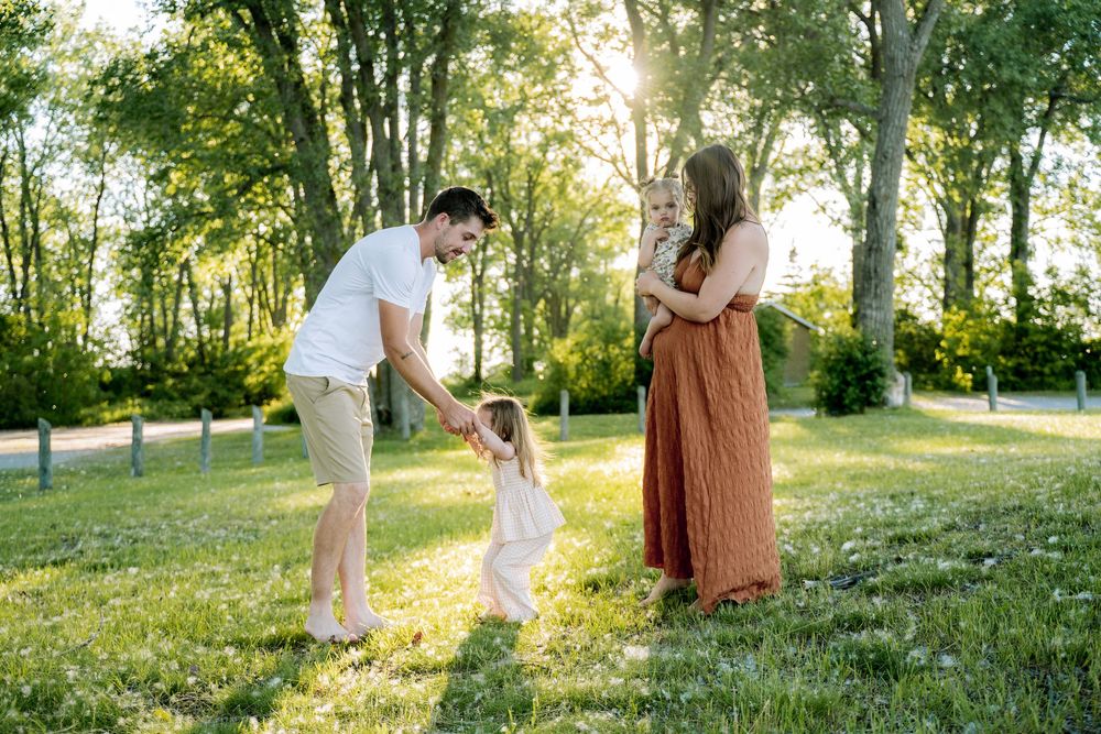Family enjoying golden hour in a park, father holding toddler's hands while pregnant mother watches warmly.