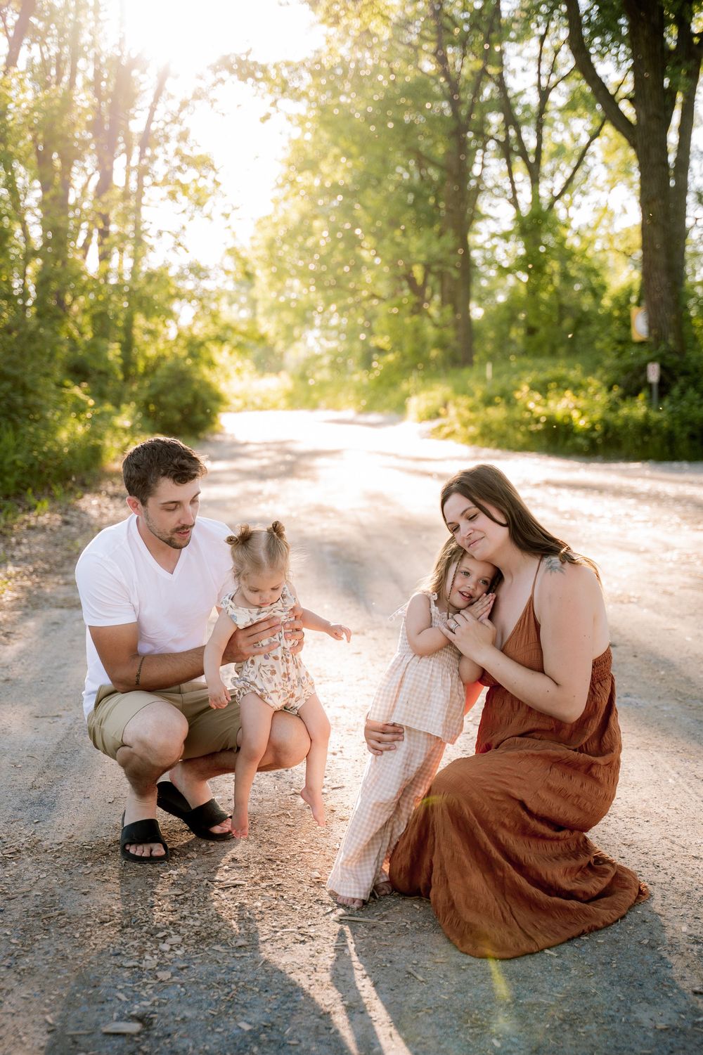 Family of four posing on a sunlit dirt road surrounded by lush green trees during golden hour outdoor photo session.