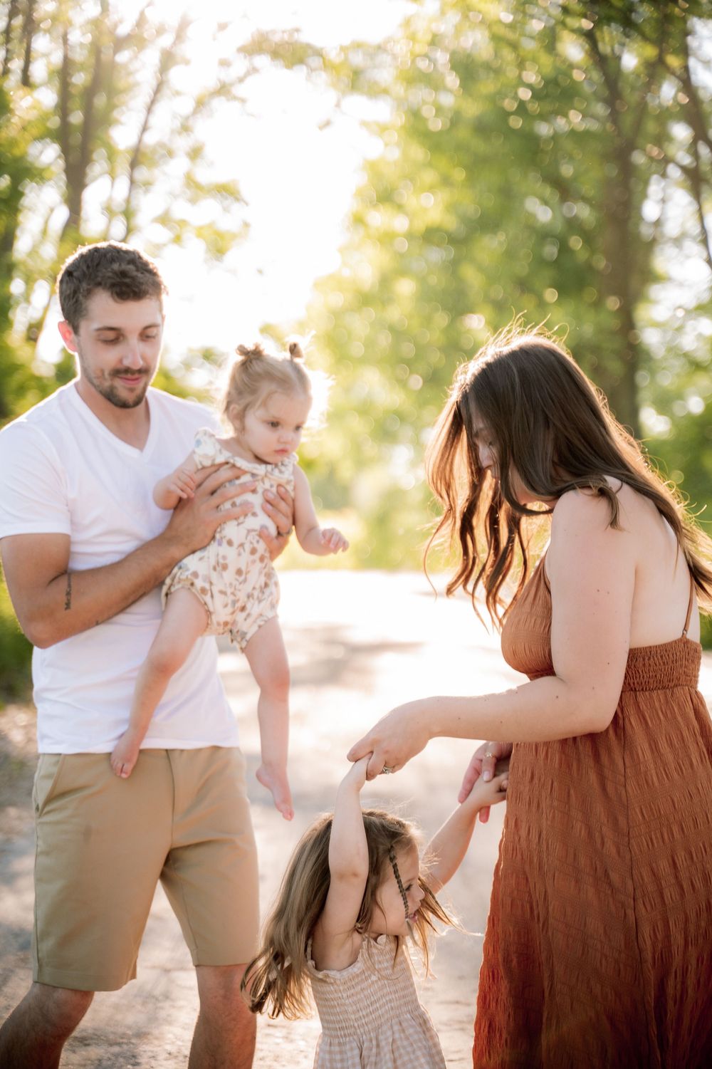 A young family enjoys a sunny outdoor moment, with dad holding a baby and mom holding a toddler's hand.
