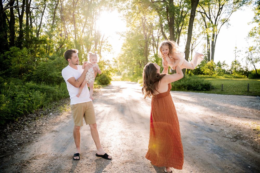 Family playing outdoors on a sunny dirt road, parents lifting children in golden hour light among lush green trees.