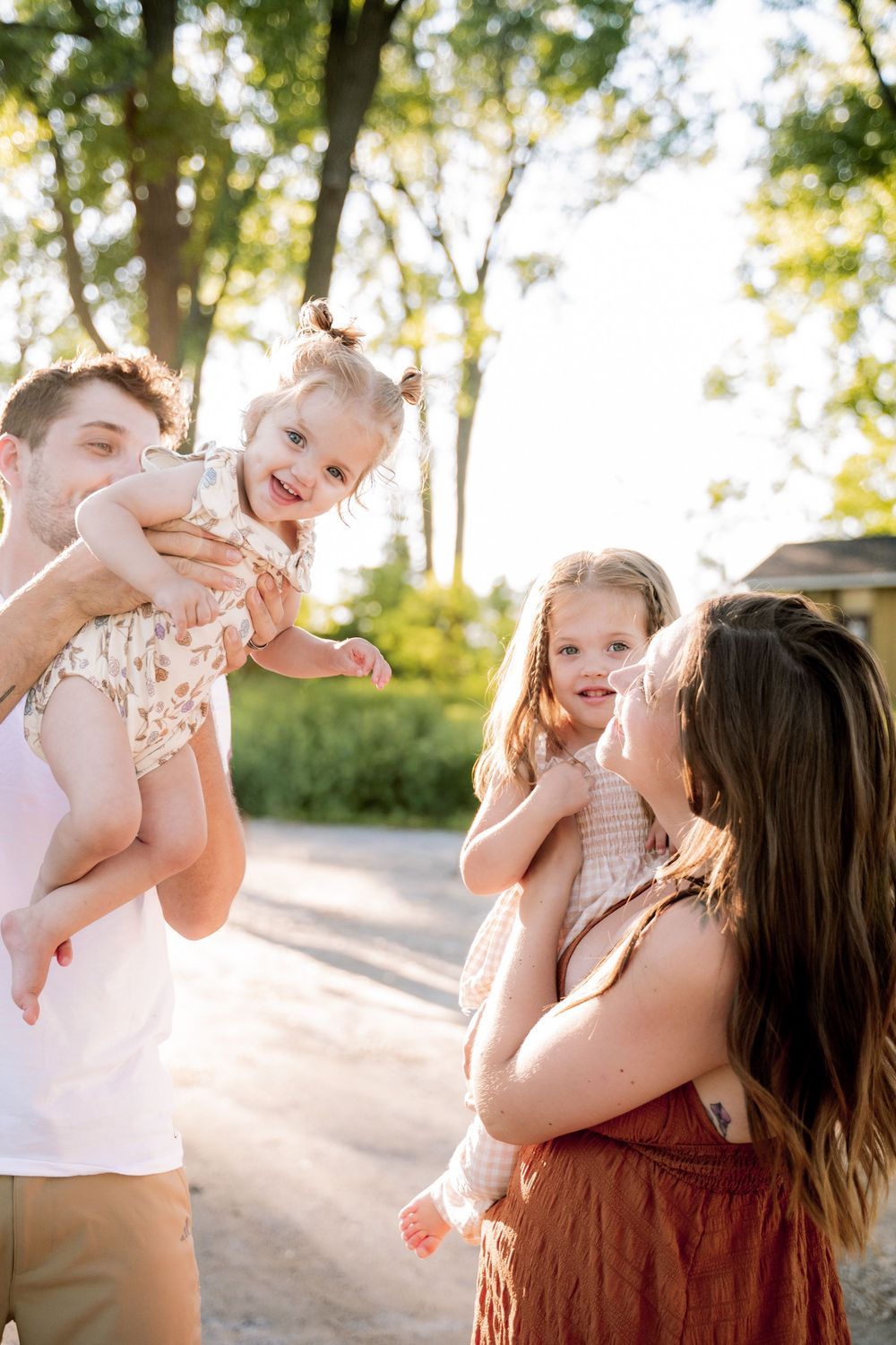 A young family enjoys a sunny outdoor moment, with parents lifting and holding two smiling toddler girls near green trees.