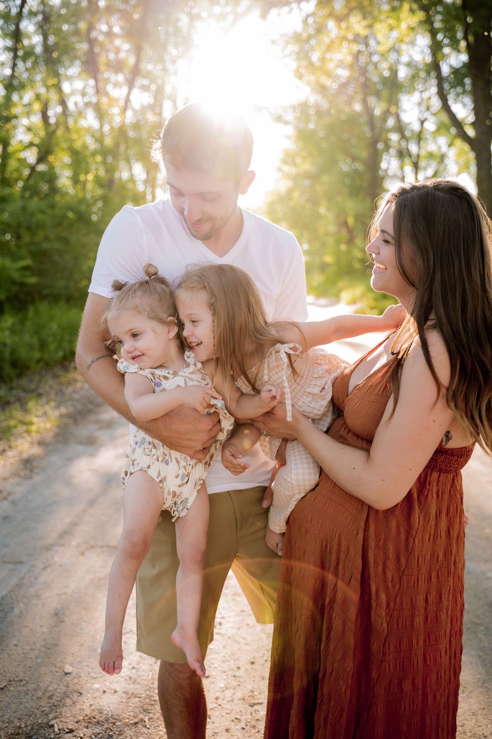 A joyful family with two young girls shares a warm moment on a sunlit dirt path surrounded by trees.