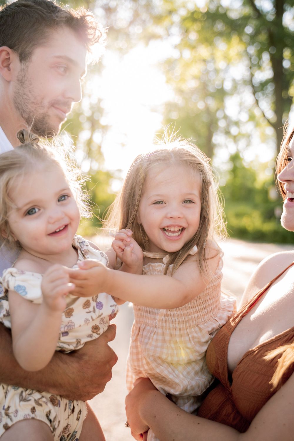 Happy family with two young girls laughing outdoors in golden sunlight, surrounded by lush green trees.