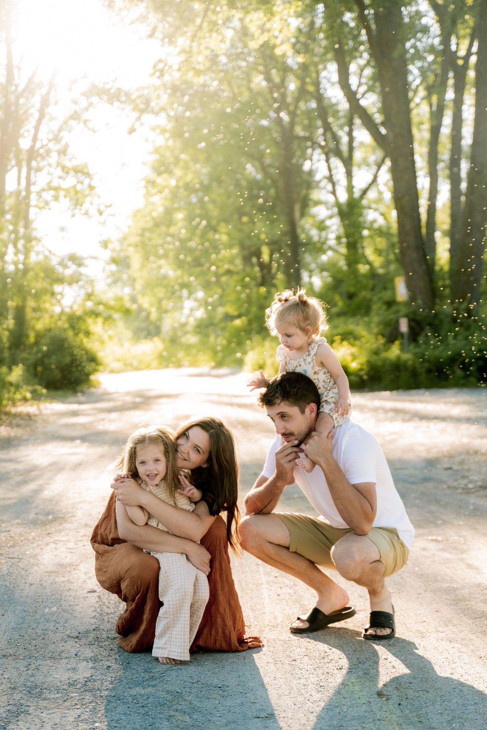 Family of four poses on a sunlit road, parents crouching with two young daughters amid lush green trees.