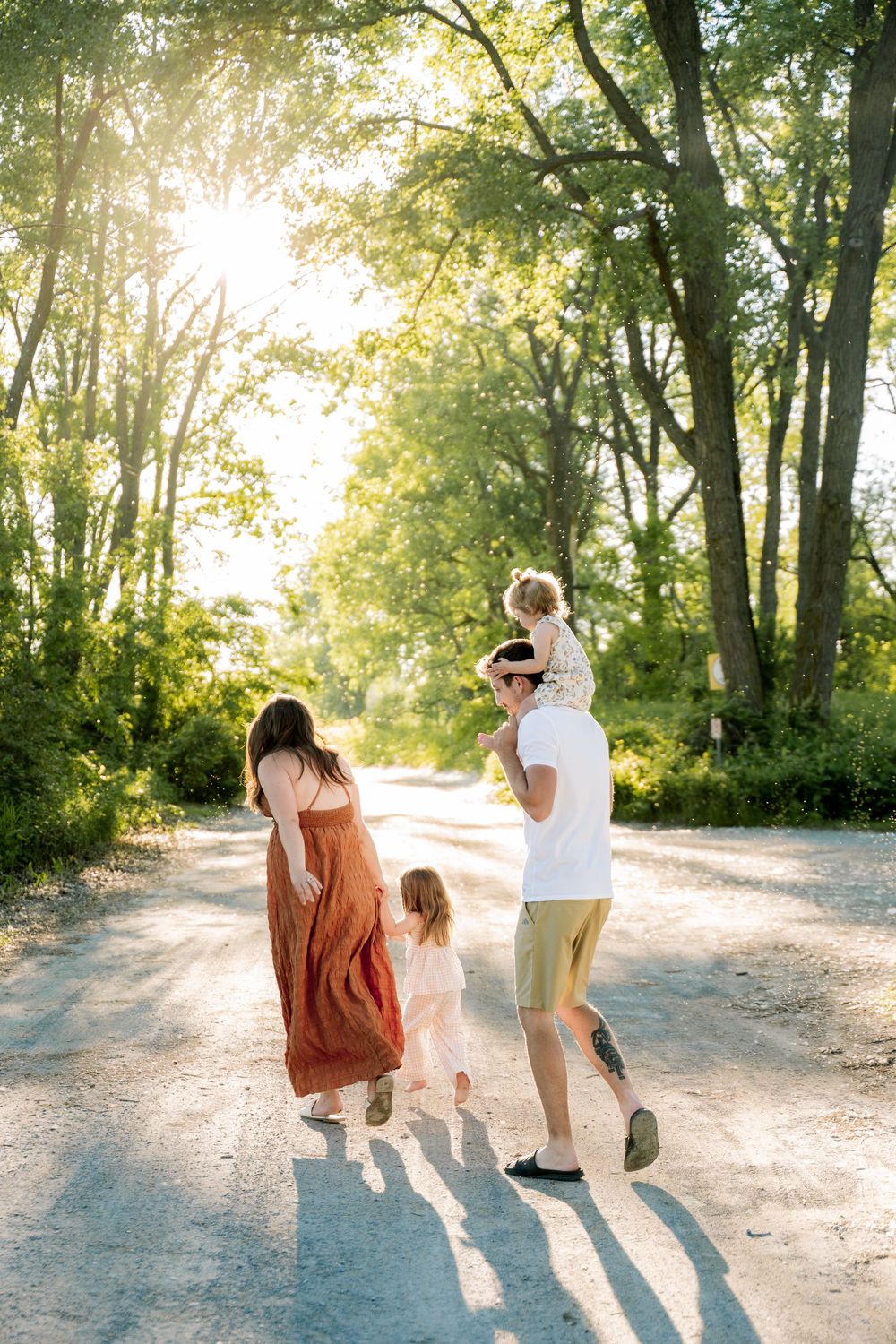 A family walks down a sunlit tree-lined path, with dad carrying a child on his shoulders and mom holding another child's hand.