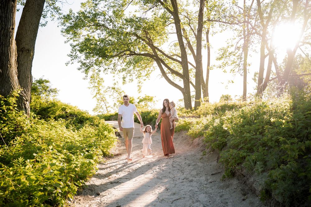 Family of three walking hand-in-hand on a sunny sandy path surrounded by lush green trees and plants.