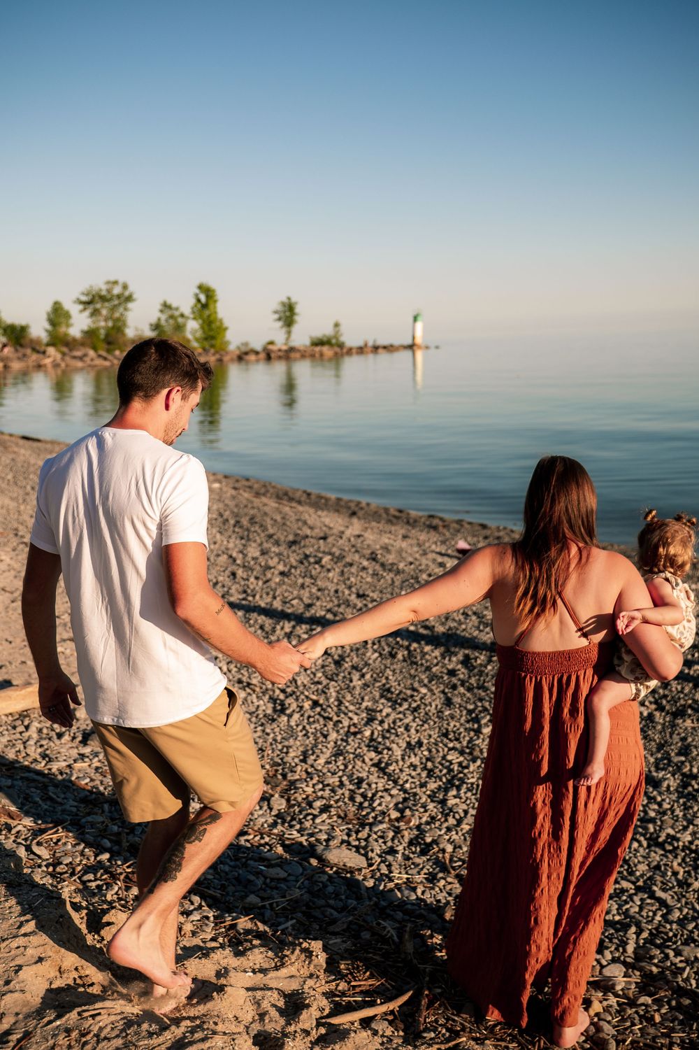 Family of three walking hand-in-hand on a rocky beach at sunset, with calm water and trees in the background.