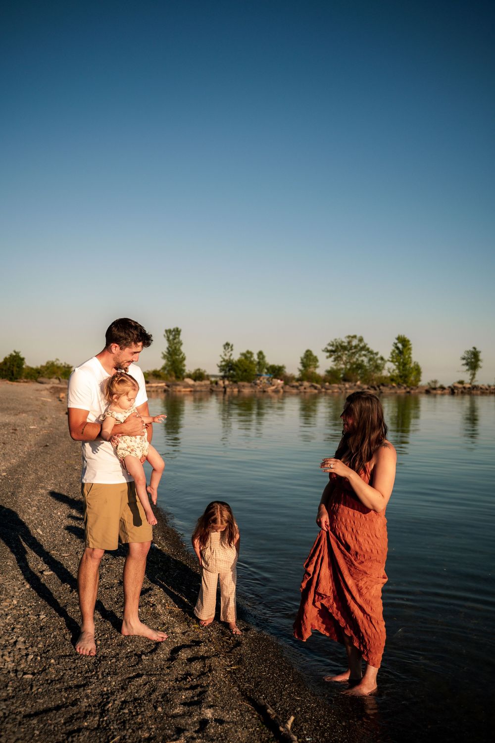 Family of four enjoys a sunny lakeside moment, parents with two young children standing at the water's edge.