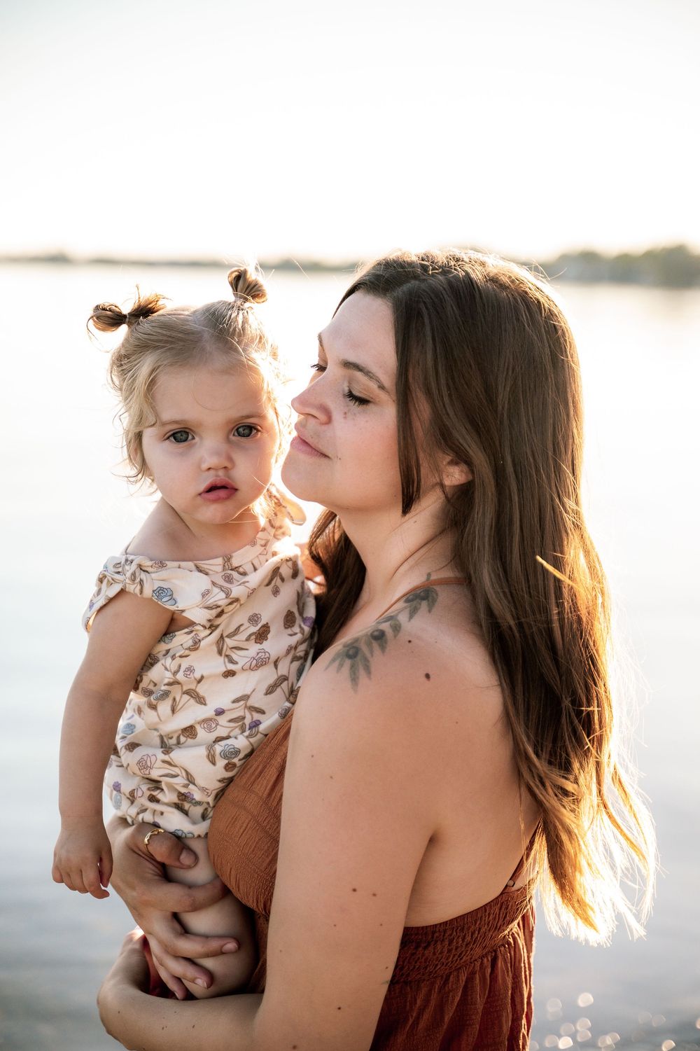 A young mother holds her toddler daughter near a lake at golden hour sunset.
