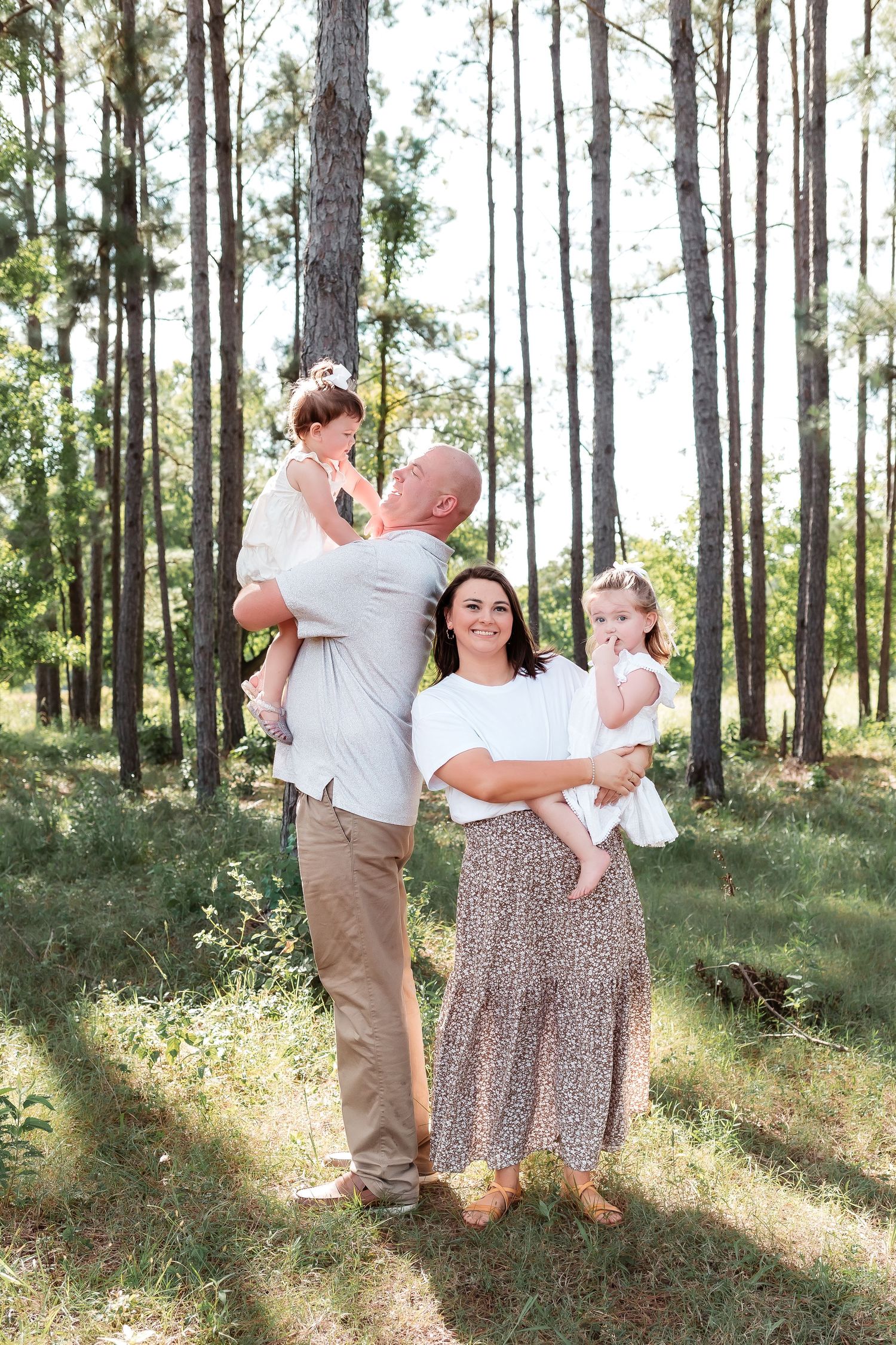 Family of four posing outdoors in a pine forest, parents holding two young children, wearing neutral tones.