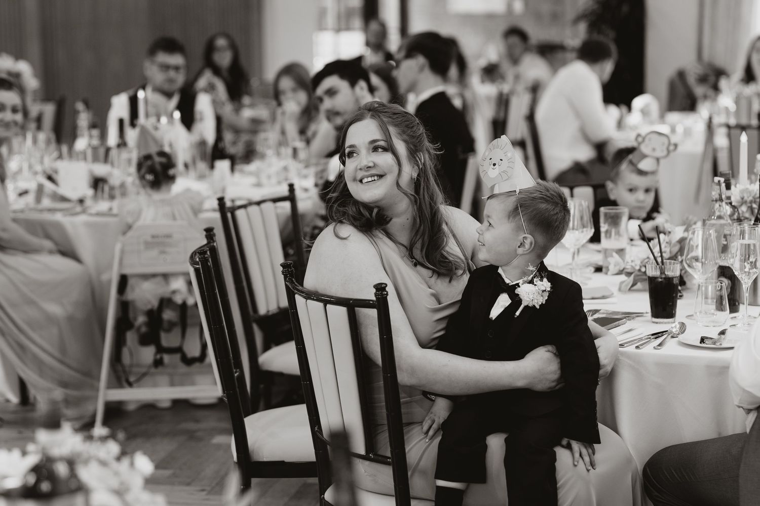 Black and white wedding reception photo of smiling bride and groom seated at decorated table with guests.