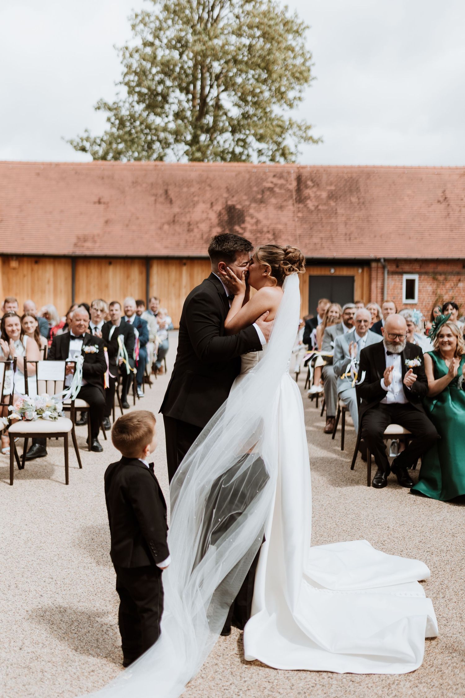 Bride and groom share first kiss at outdoor barn wedding ceremony while young ring bearer stands nearby.