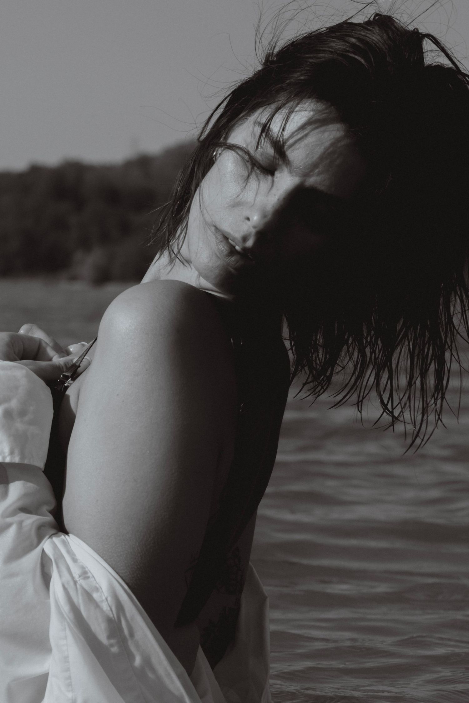 Black and white photo of a woman with wet hair near water, wearing a white dress, eyes closed in a serene pose.