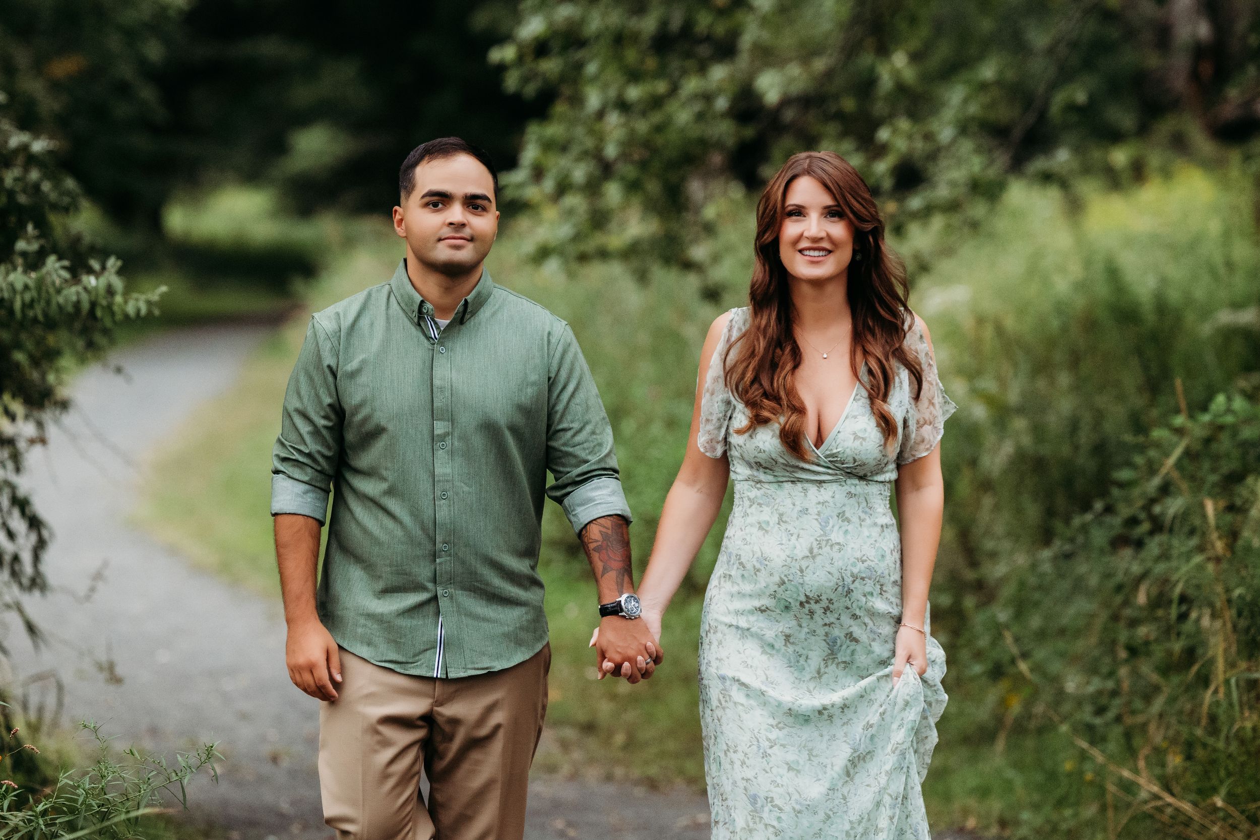 Couple holding hands walking on a scenic path surrounded by lush green trees during an outdoor engagement session.