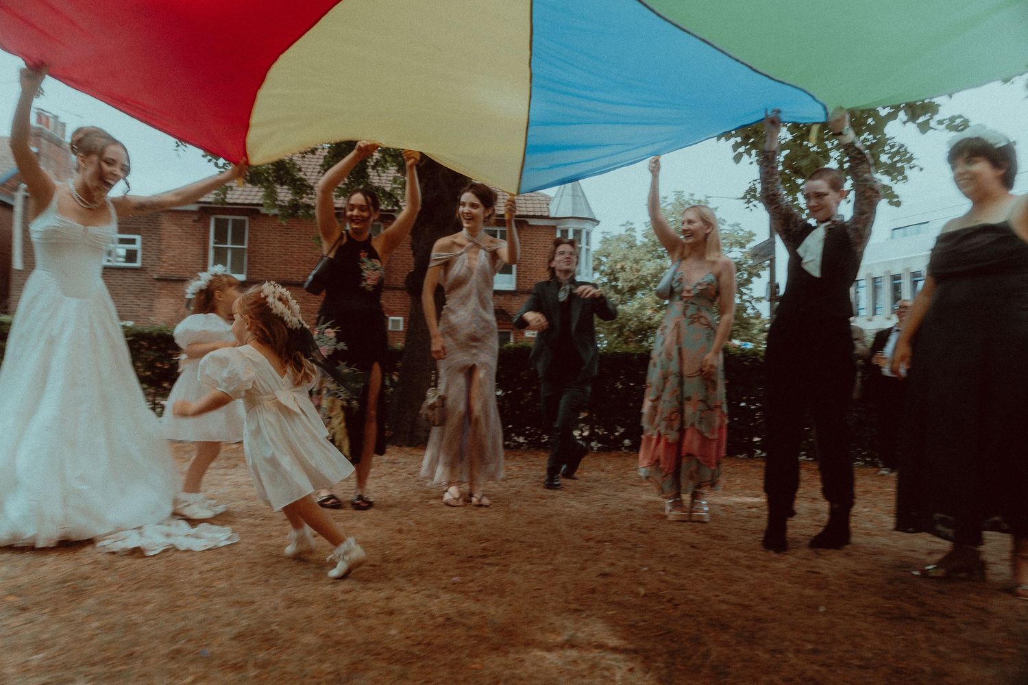 Wedding guests dance joyfully under a colorful rainbow parachute outdoors, with a bride in white gown celebrating.