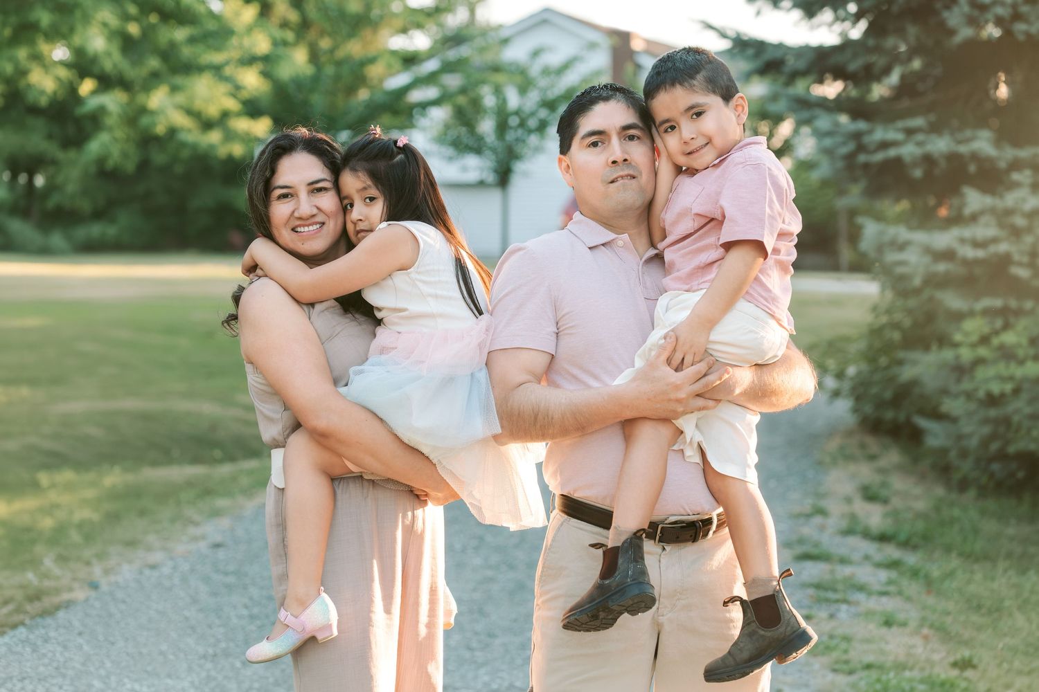 A happy family of four poses outdoors, parents holding two young children, smiling warmly in a lush green setting.