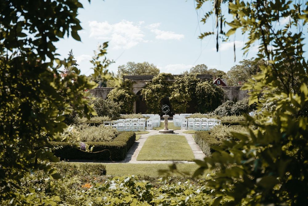Elegant outdoor wedding ceremony setup in a formal garden with white chairs, manicured hedges, and stone architecture.