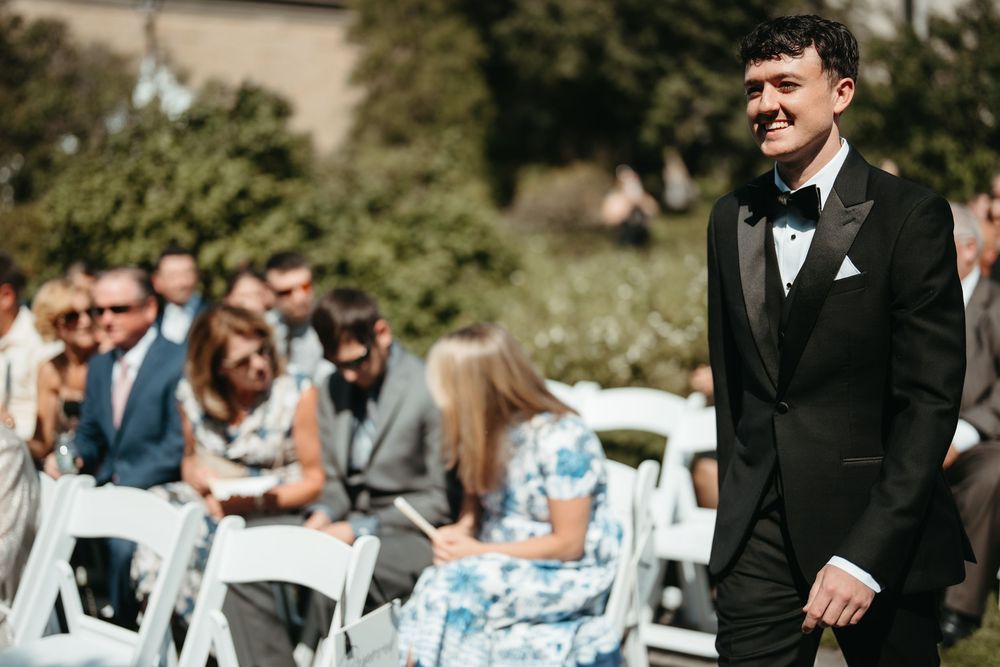 Groom in black tuxedo smiling joyfully at outdoor wedding ceremony with seated guests in background.