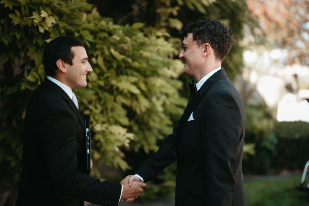 Two men in black suits shaking hands outdoors at a formal event, surrounded by lush green trees.
