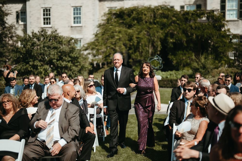 Couple walks down outdoor wedding aisle surrounded by seated guests on sunny day at elegant venue.