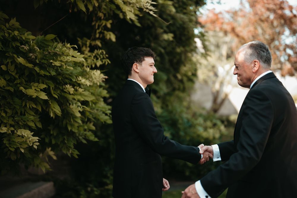 Two men in formal black suits shaking hands outdoors near lush green hedges at a wedding event.