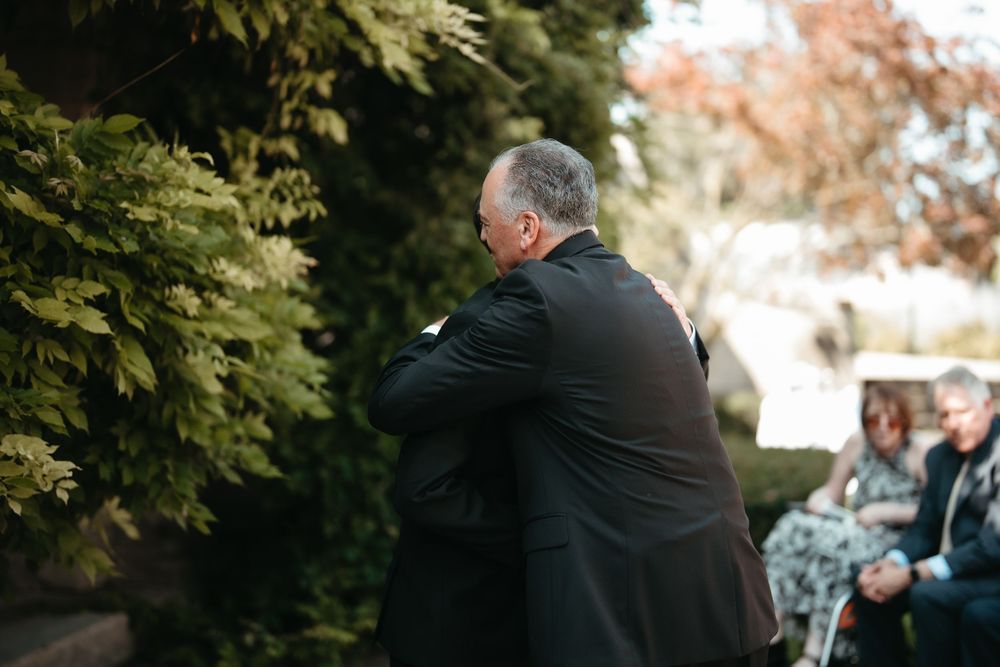 Two men in black suits share an emotional hug outdoors at a formal gathering with guests seated nearby.