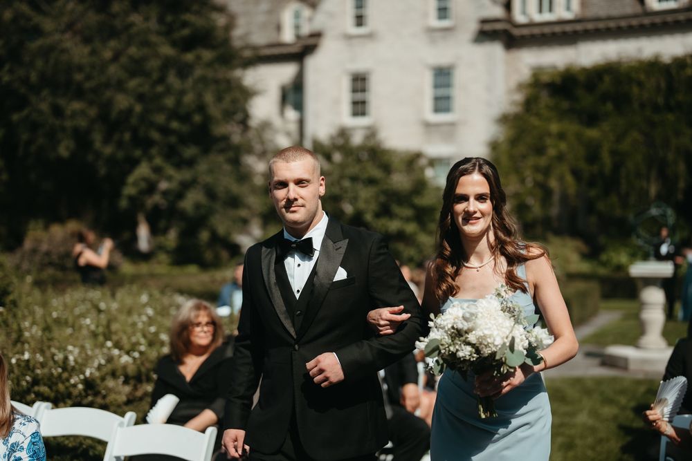 Groomsman escorts bridesmaid in light blue dress down outdoor wedding ceremony aisle at elegant estate.