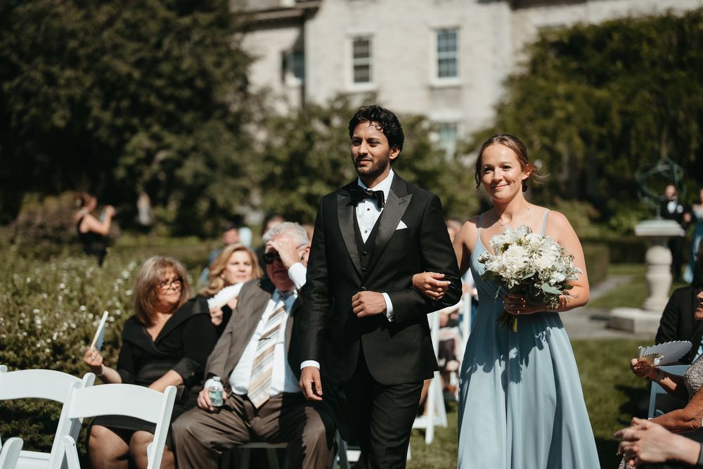 A groomsman in a black tuxedo escorts a bridesmaid in a blue gown down the aisle at an outdoor wedding ceremony.