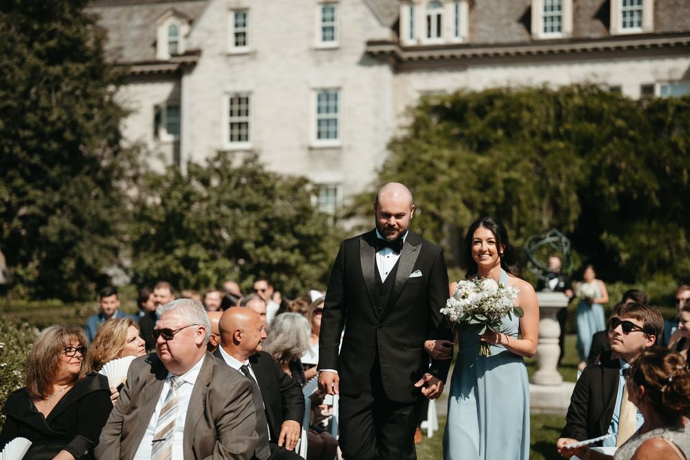 Groomsman walks bridesmaid holding white bouquet down sunlit outdoor wedding aisle at historic venue.