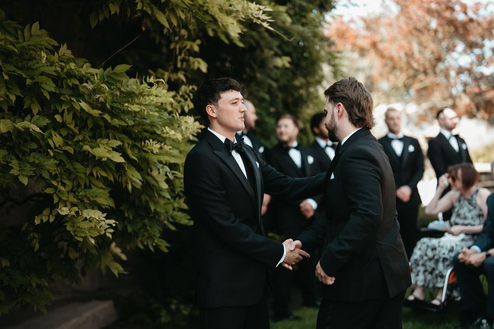Two groomsmen in black tuxedos share a handshake at an outdoor wedding ceremony near lush greenery.